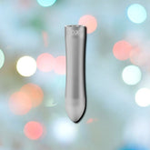 A sleek, cylindrical metallic object with a slightly tapered end, positioned vertically against a multicolored, blurred background featuring soft circular bokeh lights in shades of white, green, orange, and pink. The Doxy Bullet Vibrator - Silver by Doxy promises intense vibrations perfect for precision play.